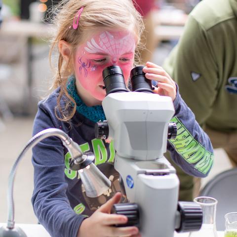 A girl with face painted like a butterfly looks into a microscope. 